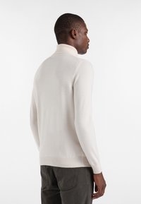 Valkoinen pooloneule, jossa on ribattu kaulus ja hihansuut, tiukka malli, yhdistettynä harmaisiin kuvioituihin housuihin. Sujuva texture, minimaalinen sauma.