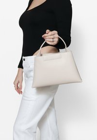 Beige leren handtas met een gestructureerde vorm en een korte, gebogen handgreep. Glad oppervlak en minimalistisch ontwerp zonder zichtbare hardware.