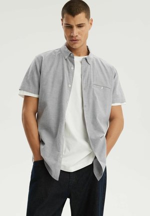 Młody mężczyzna ubrany w jasnoszarą, krótkawą koszulę na guziki na białą koszulkę oraz ciemne spodnie, stoi z rękami w kieszeniach.