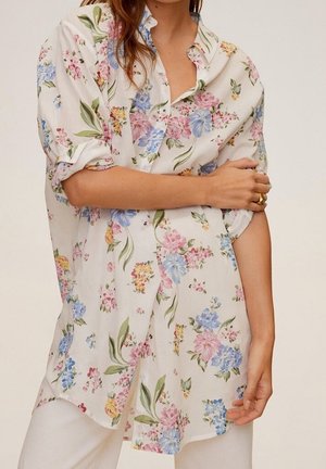 Kobieta w białej koszuli zapinanej na guziki z niebieskimi, różowymi i żółtymi kwiatami, z podwiniętymi rękawami, w połączeniu z białymi spodniami.