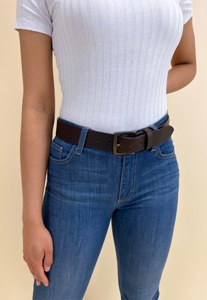 Femme portant un haut blanc côtelé à manches courtes, rentré dans un jean bleu avec une ceinture en cuir marron foncé avec une boucle en laiton.