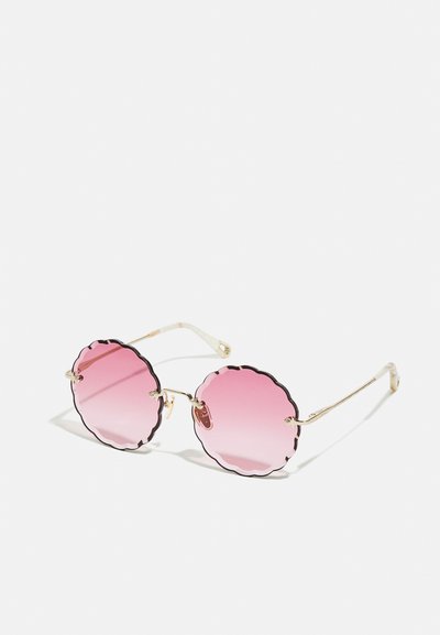 Okrugle sunčane naočale s valovitim ružičastim gradijentnim staklima i tankim zlatnim metalnim okvirima na bijeloj pozadini.
