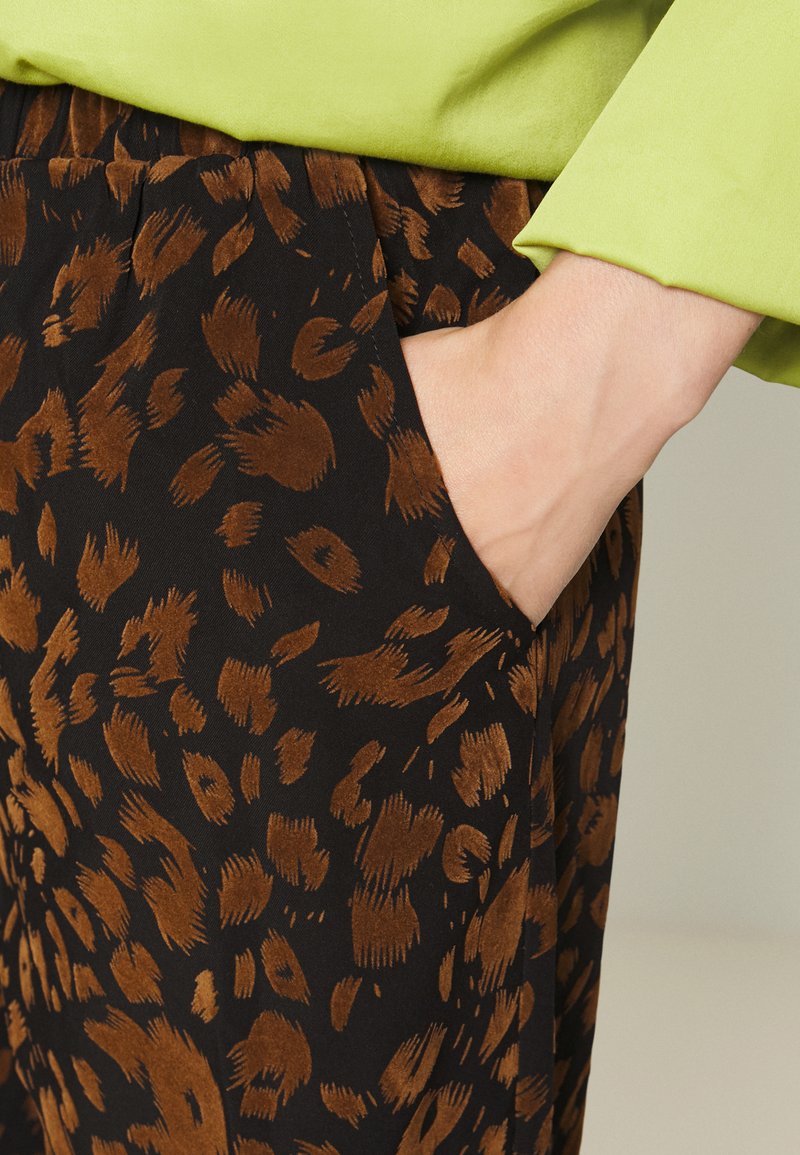 Schwarzer Stoff mit einem braunen Leopardenmuster. Eine Hand befindet sich in der Seitentasche der Hose. Der Stoff wirkt weich.