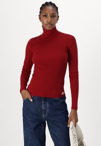 Piros bordás garbós pulóver hosszú ujjakkal és feszes szabással. Az alján egy kis logós címke található. Sötét denim farmernadrággal párosítva.