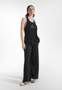 Sort ærmeløs top med "Re-pose" grafik, kombineret med løse sorte bukser. Stoffet ser blødt og glat ud med en afslappet pasform. Lys baggrund.