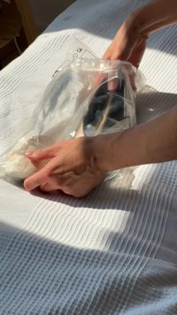Mani che posano scarpe nere avvolte in plastica trasparente su una superficie di tessuto bianco texturizzato, illuminate dalla luce solare laterale.