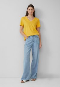 T-shirt jaune à manches courtes et col en V associé à un jean large bleu clair, avec une coupe décontractée et une texture subtile, porté avec des sandales claires.