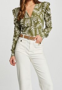 Blouse verte avec motif de feuilles de palmier blanches, manches bouffantes et encolure en V, associée à un pantalon blanc taille haute et une ceinture marron clair.