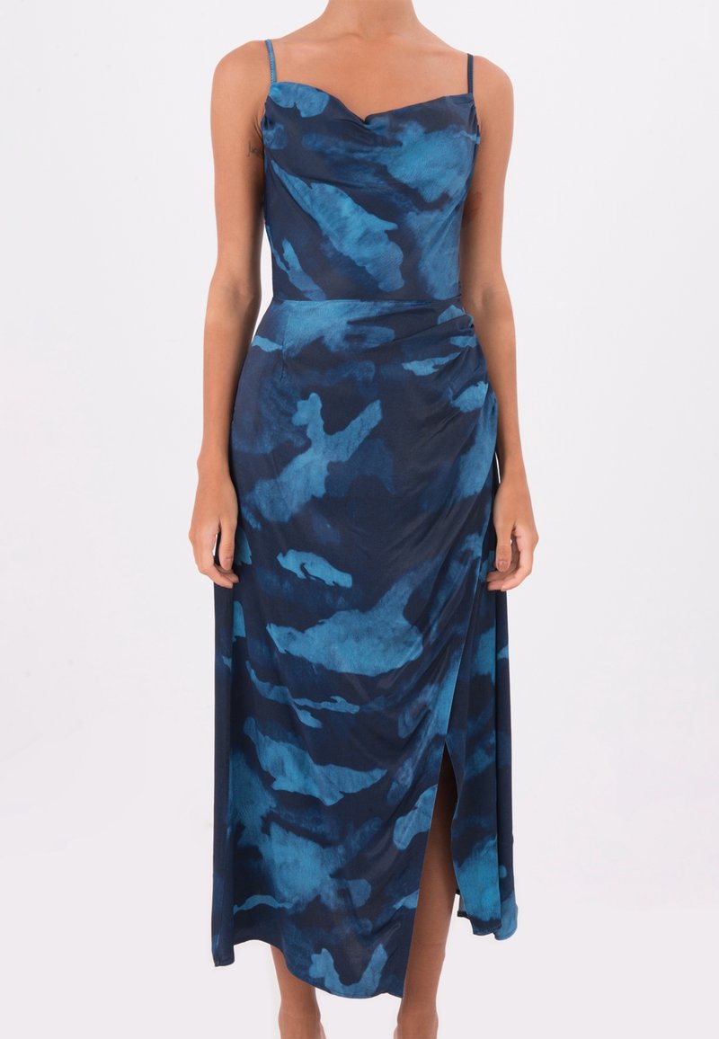 Femme portant une robe sans manches bleue et bleu marine à motifs, avec de fines bretelles et une fente à l'avant, debout devant un fond blanc uni.