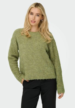 Groene gebreide trui met een relaxte pasvorm, ronde hals en lange mouwen. Zachte textuur; de zoom is lichtjes rafelig. Gecombineerd met zwarte broek.