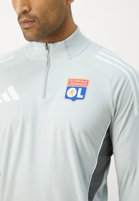 Lysegrå treningsjakke med høy krage, glidelås foran og sidelister. Har et Olympique Lyonnais-logo på brystet.