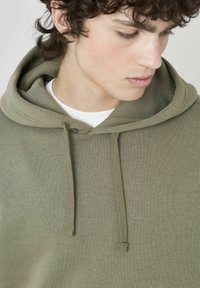 Hoodie vert olive en tissu doux, avec une capuche à cordons, une texture côtelée et une coupe décontractée. Design uni sans motifs.