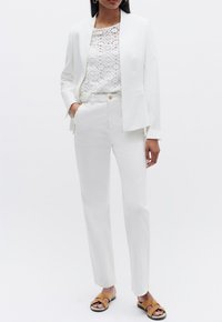 Blazer bianco su un top in pizzo bianco, abbinato a pantaloni bianchi a gamba dritta e sandali beige. Design pulito con toni neutri.
