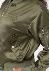 Olijfgroene bomberjack van satijn, met geborduurde bloemenmotieven, ribgebreide manchetten, twee zijvakken en een ritsvak op de mouw.