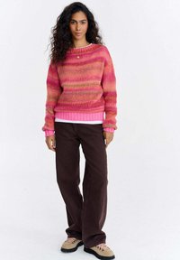 Kobieta stojąca, ubrana w różowo-pomarańczowy sweter w paski, ciemnobrązowe spodnie, beżowe buty trekkingowe, z długimi kręconymi ciemnymi włosami, na jednolitym białym tle.