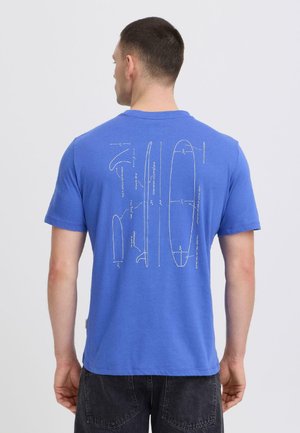 Hombre con una camiseta azul con dibujos técnicos blancos y medidas de una tabla de surf en la espalda, combinado con pantalones negros.