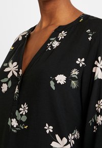 Blouse florale noire présentant des motifs de fleurs blanches et vertes, un décolleté en V et des manches amples en tissu léger.