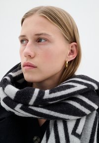 Sciarpa di lana a righe bianche e nere avvolta attorno al collo, con una texture liscia. Orecchino a cerchio dorato visibile sull'orecchio del modello.