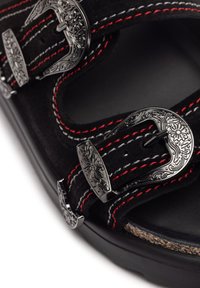 Sandal in suede nero con ornate fibbie argentate, cuciture rosse e bianche, un fondo testurizzato e una suola nera robusta.