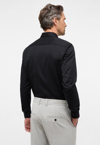 Camicia nera con colletto classico, maniche lunghe e polsini abbottonati, abbinata a un paio di pantaloni grigi chiari con texture e tasche posteriori.