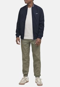 Giubbotto bomber navy con collo a costine, maglione crema a girocollo, pantaloni jogger verde oliva e sneaker beige con dettagli chiari e suole in gomma.