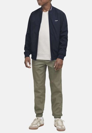 Giubbotto bomber navy con collo a costine, maglione crema a girocollo, pantaloni jogger verde oliva e sneaker beige con dettagli chiari e suole in gomma.