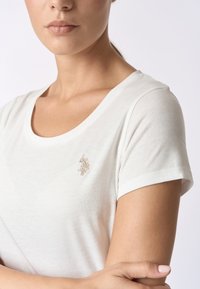 T-shirt bianco a maniche corte con scollatura rotonda. Presenta un piccolo logo metallico ricamato sull'area del petto sinistro. Tessuto dal tocco liscio.