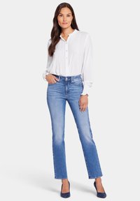 Blusa blanca con plisados y botones en el frente, combinada con jeans rectos de color azul claro. La modelo lleva tacones azul marino y se apoya con una mano en la cadera.