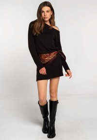 Suéter de punto negro con un diseño off-shoulder, combinado con una faja de cintura en patrones burdeos y dorado, y botas de cuero negras hasta la rodilla.