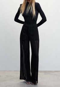 Femme portant un haut ajusté à manches longues noir et un pantalon noir large et transparent, se tenant les mains derrière le dos contre un fond uni.