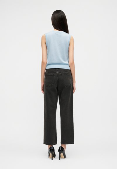 Frau mit langem, dunklem Haar, die von hinten zu sehen ist, trägt ein ärmelloses, hellblaues Oberteil, schwarze High-Waist-Jeans und schwarze High-Heel-Schuhe.