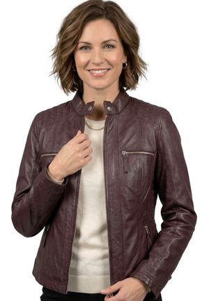Smiling woman with short wavy hair wearing a burgundy leather jacket, beige sweater, and gold chain necklace on a white background.