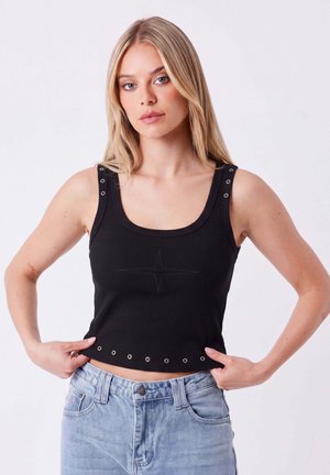 Joven mujer con cabello largo y rubio que lleva una camiseta sin mangas negra con ojales metálicos y vaqueros azul claro, posando con las manos en las caderas.