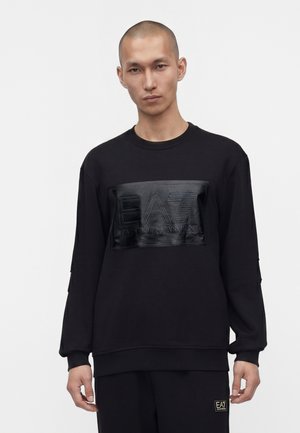 Homme à la tête rasée portant un sweat-shirt noir à manches longues avec le logo EA7 Emporio Armani en relief sur la poitrine sur fond blanc.