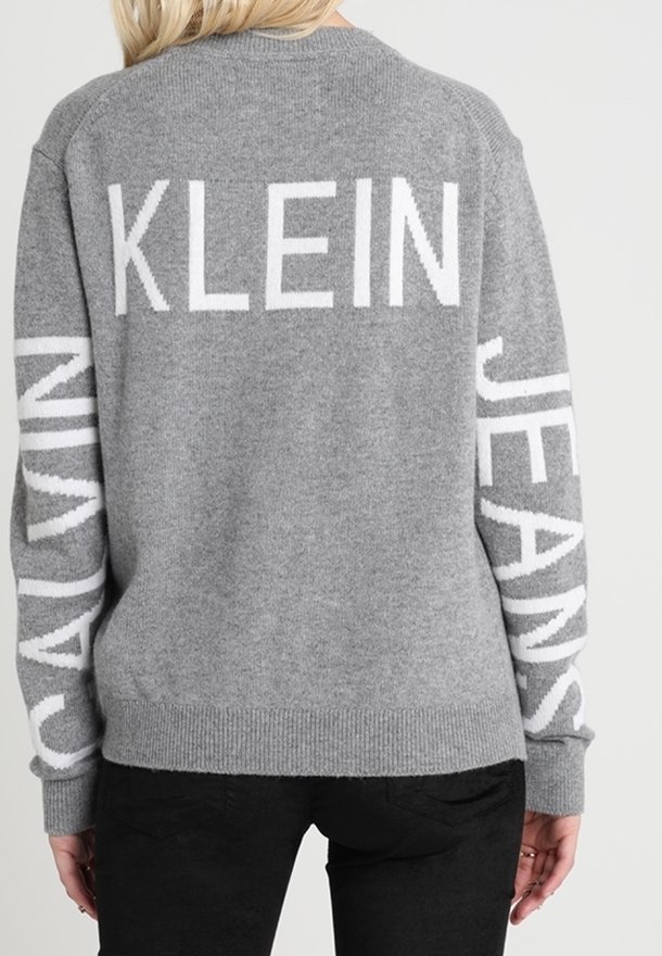 En blond kvinna som bär en grå tröja med "KLEIN" på baksidan, "CALVIN" på vänster ärm och "JEANS" på höger ärm, i kombination med svarta byxor.