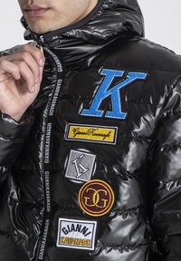 Chaqueta acolchada negra con un acabado brillante, que presenta una variedad de parches bordados coloridos y una cremallera con detalles de marca en el cuello.