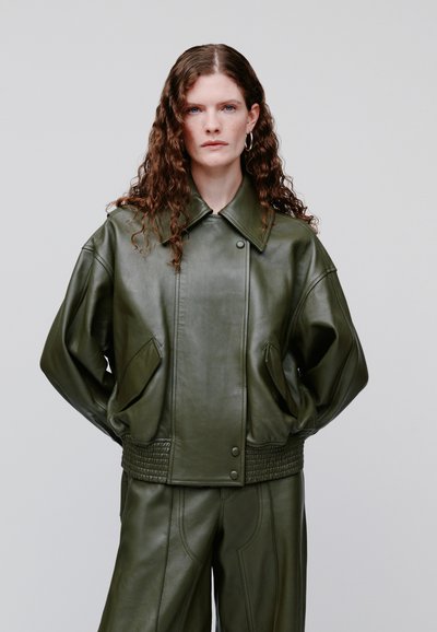 Femeie cu păr creț purtând o geacă supradimensionată de piele verde închis și pantaloni asortați, stând în fața unui fundal gri simplu.