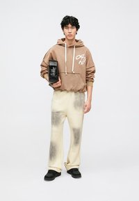 Sweatshirt castanha com capuz e logótipo branco, calças bege com manchas escuras e bolsa preta. Modelo calça sapatos pretos. Traje simples e casual.