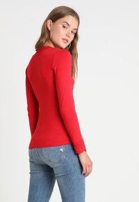 Maglietta rossa a maniche lunghe con bordo in pizzo al colletto e ai polsi, design aderente, tessuto morbido, abbinata a jeans blu chiaro.