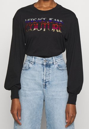 Chemise noire à manches longues avec texte coloré « Versace Jeans Couture », associée à un jean en denim bleu clair taille haute porté par une personne debout.