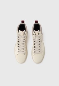 Een paar beige high-top sneakers met crèmekleurige veters, een grijze mesh kraag en rode treklussen, gezien van bovenaf op een witte achtergrond.