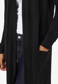 Svart lang cardigan laget av mykt stoff, med åpen front, to fremre lommer og lange ermer. Kombinert med denim underdel.