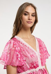 Abito rosa con motivo a fantasia, maniche a volants, rifiniture in pizzo e vita cinchata. Il tessuto è leggero e semitrasparente, con disegni floreali intricati.