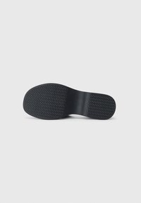 Suela de zapato negra con una superficie texturizada y un patrón de espiga. La suela tiene una forma plana con la punta redondeada.
