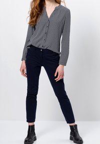 Blusa a maniche lunghe con motivo geometrico bianco e nero, abbinata a pantaloni slim-fit blu navy e stivaletti neri.