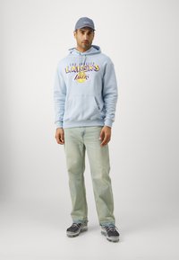 Svetlo modra kapuca z grafiko "Los Angeles Lakers", sprednja žepa in rebrasti manšeti, v kombinaciji z svetlim denimom in sivo športno obutvijo.