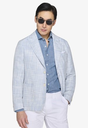 Mężczyzna w ciemnych okularach przeciwsłonecznych, niebieskiej koszuli na guziki, jasnoniebieskiej marynarce w kratę i białych spodniach, stojący na tle gładkiego tła.