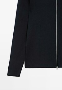 Musta ribbattu pitkähihaine vaate, jossa on pystyhopea vetoketju valkoista taustaa vasten.