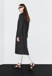 Černý dlouhý kabát z texturované tkaniny, spárovaný se světle zbarvenými přiléhavými kalhotami a tmavými sandály na podpatku, na minimalistickém pozadí.