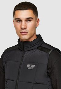 Zwarte puffer vest met een hoge kraag, ritssluiting en logo-opdruk op de borst. Beschikt over een gladde textuur en contrasterende schouderpanelen.